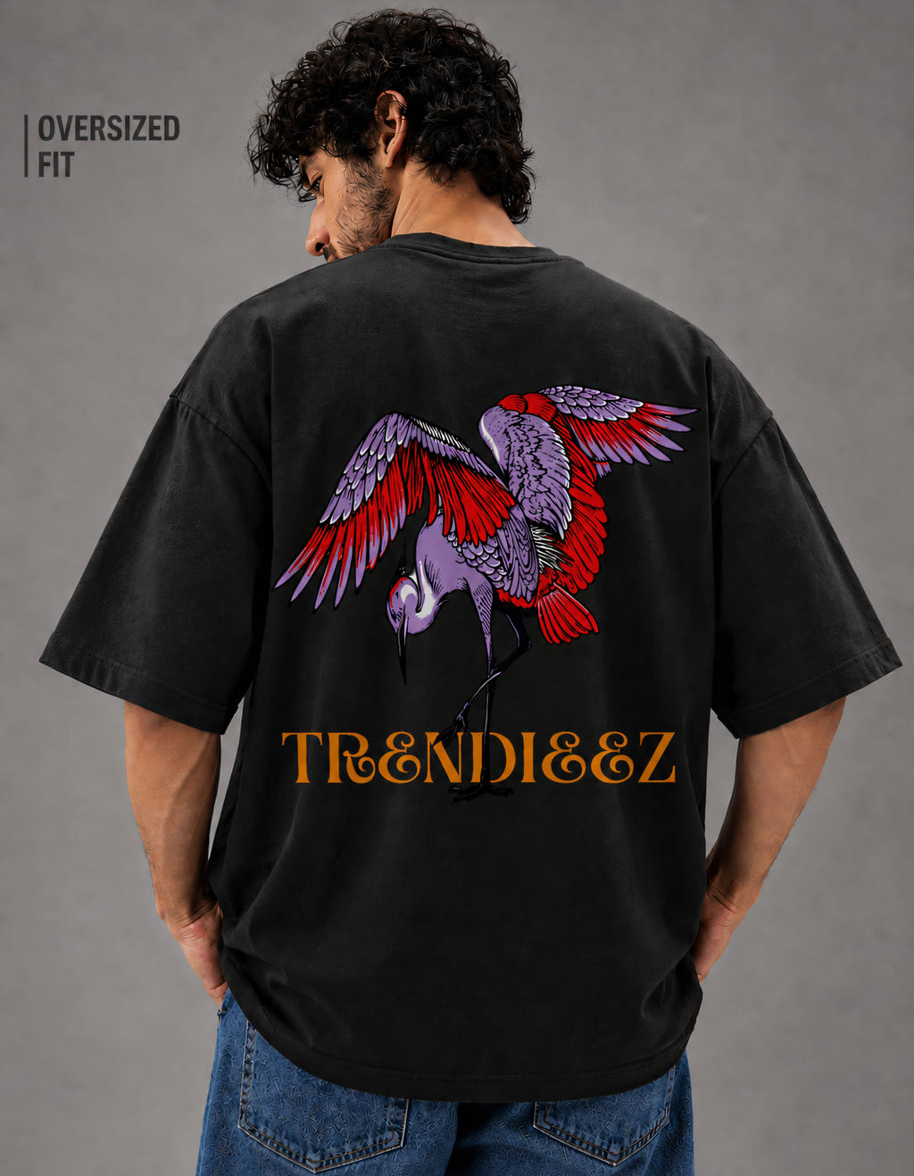 Person wearing a black oversized t-shirt with a colorful parrot design and 'TRENDIEEZ' text on the back, standing against a gray background.