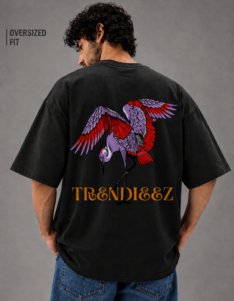 Person wearing a black oversized t-shirt with a colorful parrot design and 'TRENDIEEZ' text on the back, standing against a gray background.