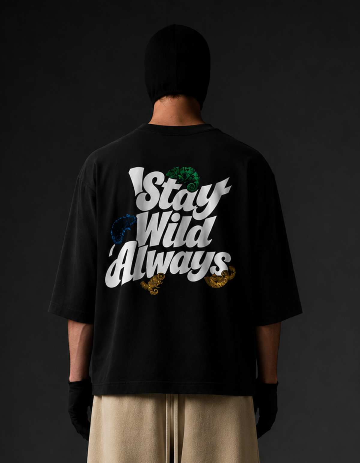 A person shown from the back wearing a black oversized T-shirt with a large graphic on the back reading 'Stay Wild Always' in white, with small green, blue, and gold accents, paired with beige pants and black gloves against a dark background.