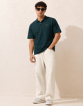 Man wearing a dark green polo shirt and white pants against a beige background