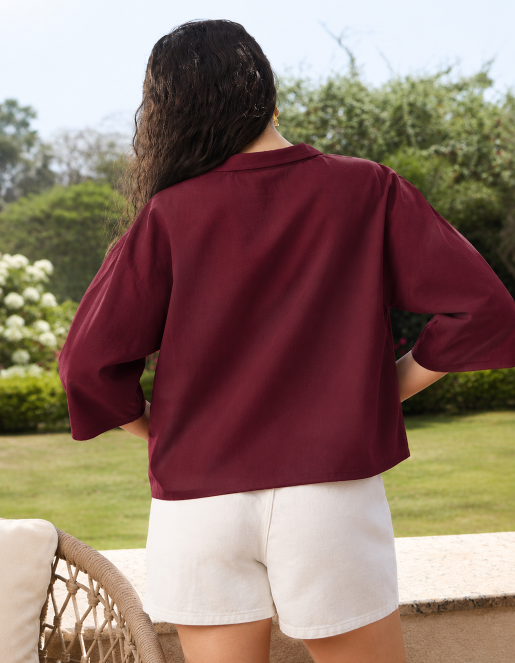 Person wearing a burgundy shirt and white shorts standing outdoors with greenery in the background
