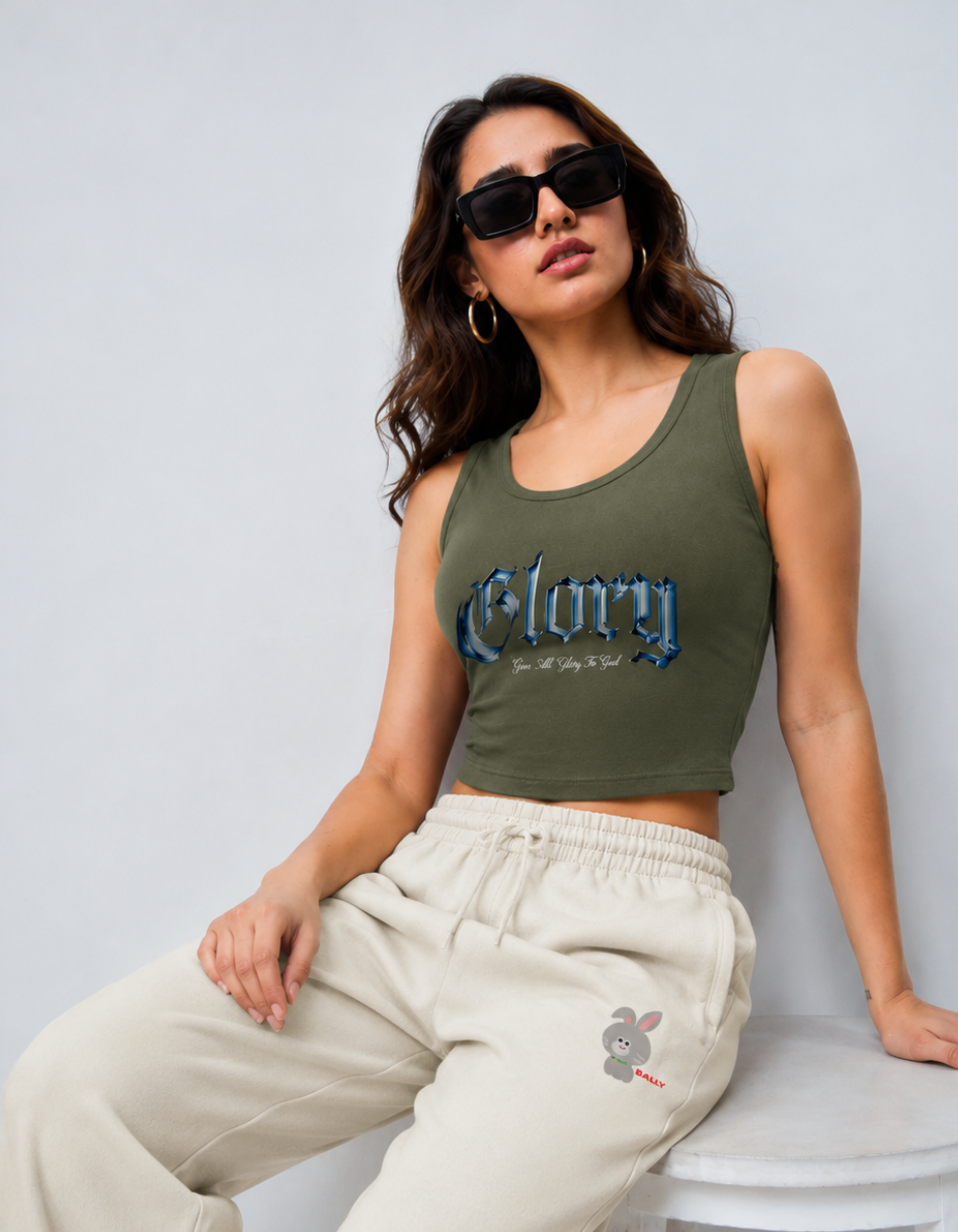 Woman wearing a green tank top with 'Glory' printed on it, sitting on a white stool against a light gray background.
