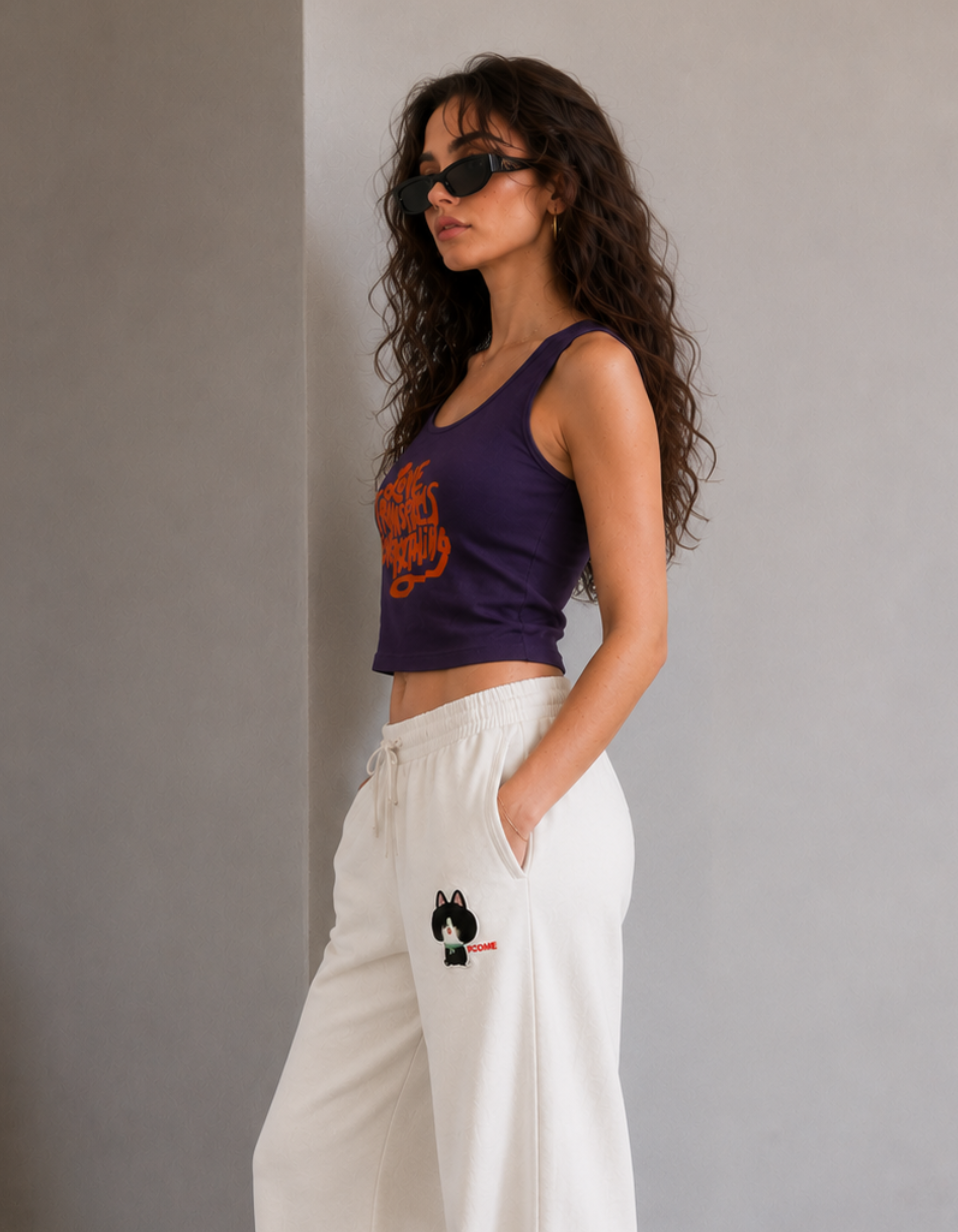 Woman wearing a purple tank top with orange design and white pants with dog embroidery against a gray background