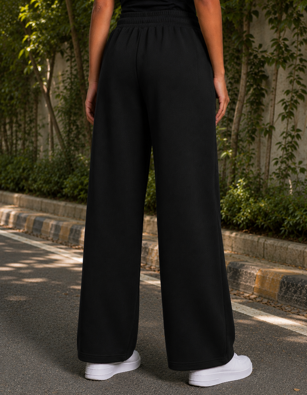 Person wearing black wide-leg pants and white sneakers standing outdoors with greenery in the background