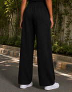 Person wearing black wide-leg pants and white sneakers standing outdoors with greenery in the background