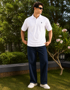 Man wearing a white polo shirt and navy pants standing outdoors with greenery in the background