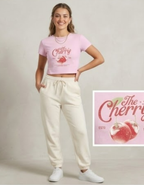 Woman wearing a pink t-shirt with 'The Cherry' design and white pants, standing against a plain background.