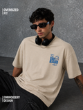 Man wearing a beige t-shirt with embroidery design, sitting against a gray background.